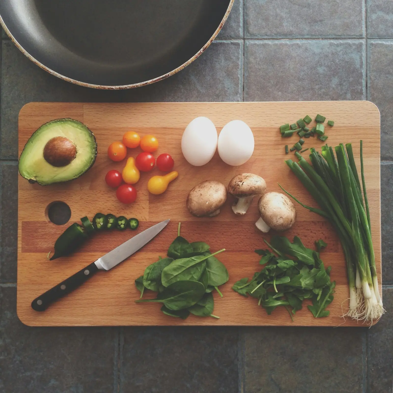 Eggs, avocado, tomatoes, mushrooms, spring onions and a knife laid out on a chopping board
