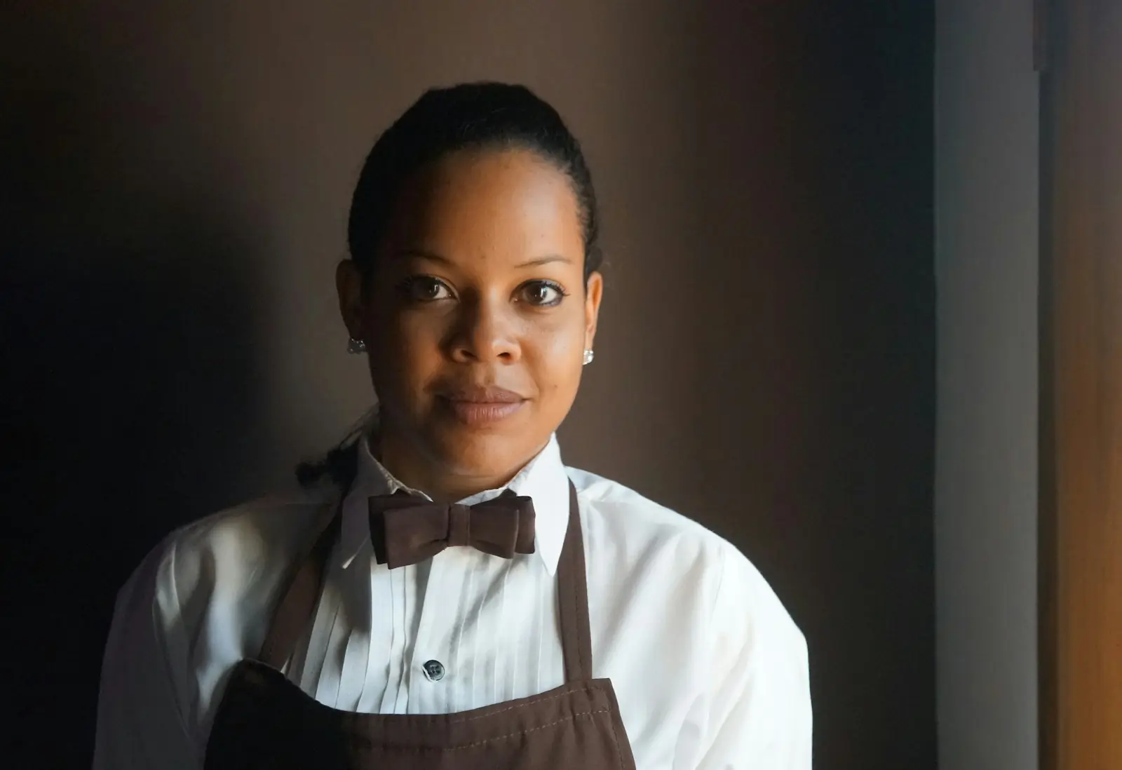 Portrait of Nadia Okafor, hair pulled back, in a white shirt, bow tie and brown apron
