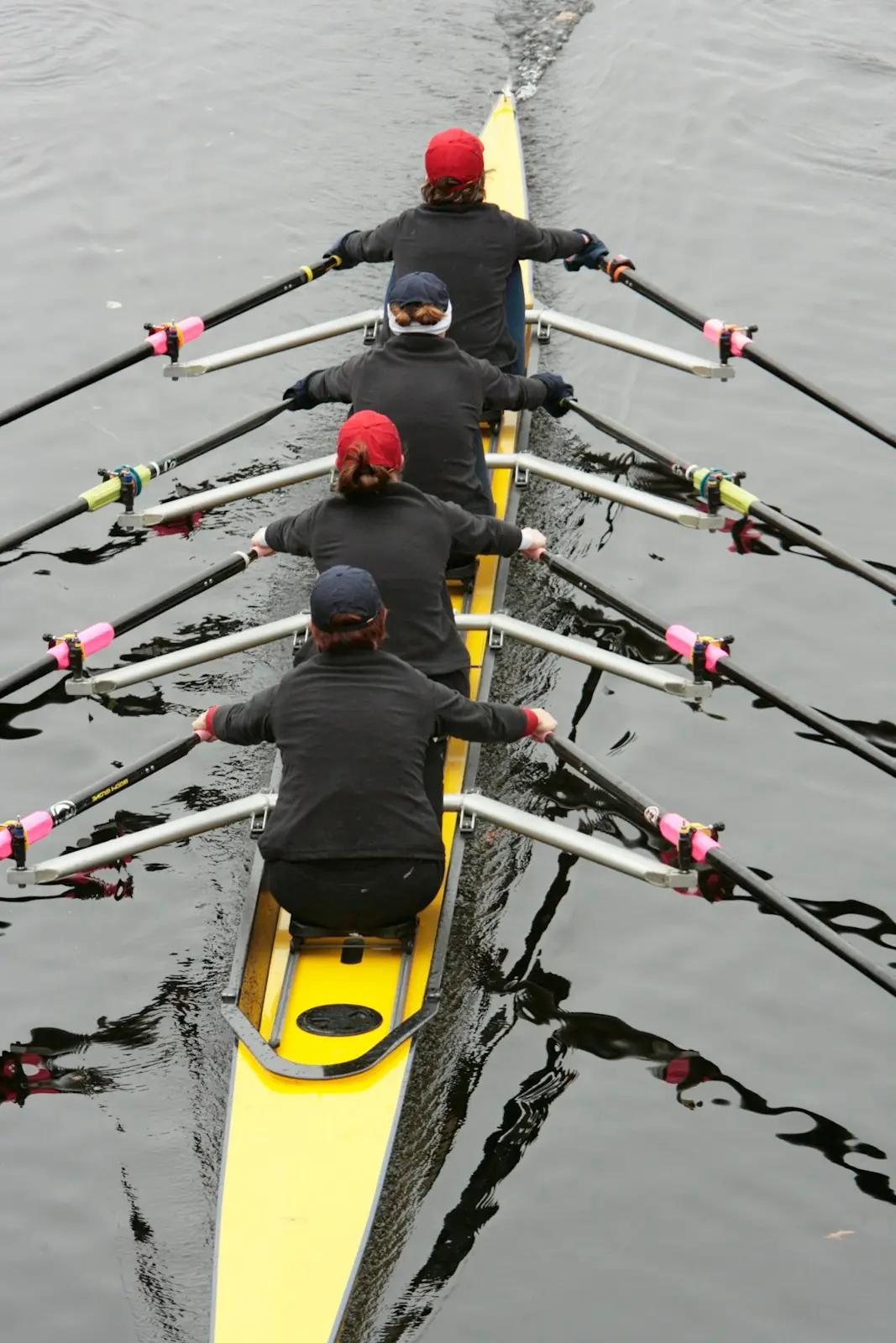 From behind the stroke seat: rowers in dark tops pulling together, oars with pink and white collars spread over the water