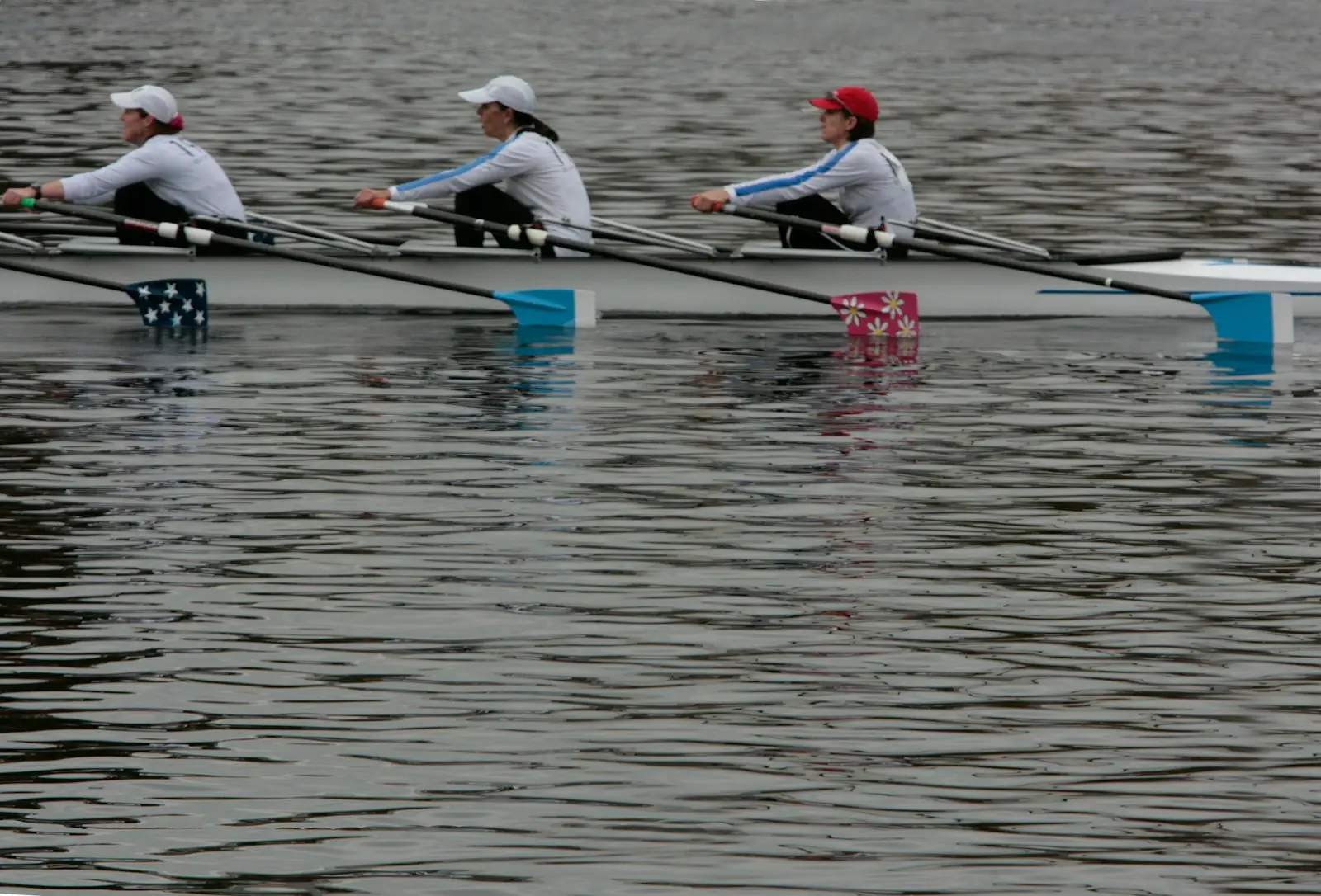 Three rowers in caps and light tops sculling side by side on a calm grey river