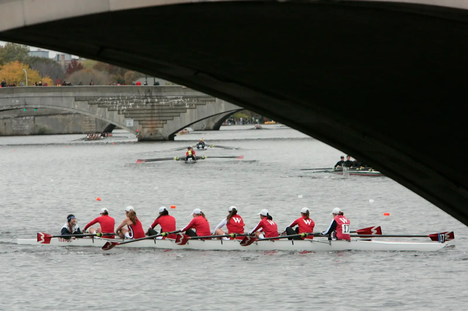 A junior crew in red shirts racing an eight under a stone bridge, a coach's launch behind