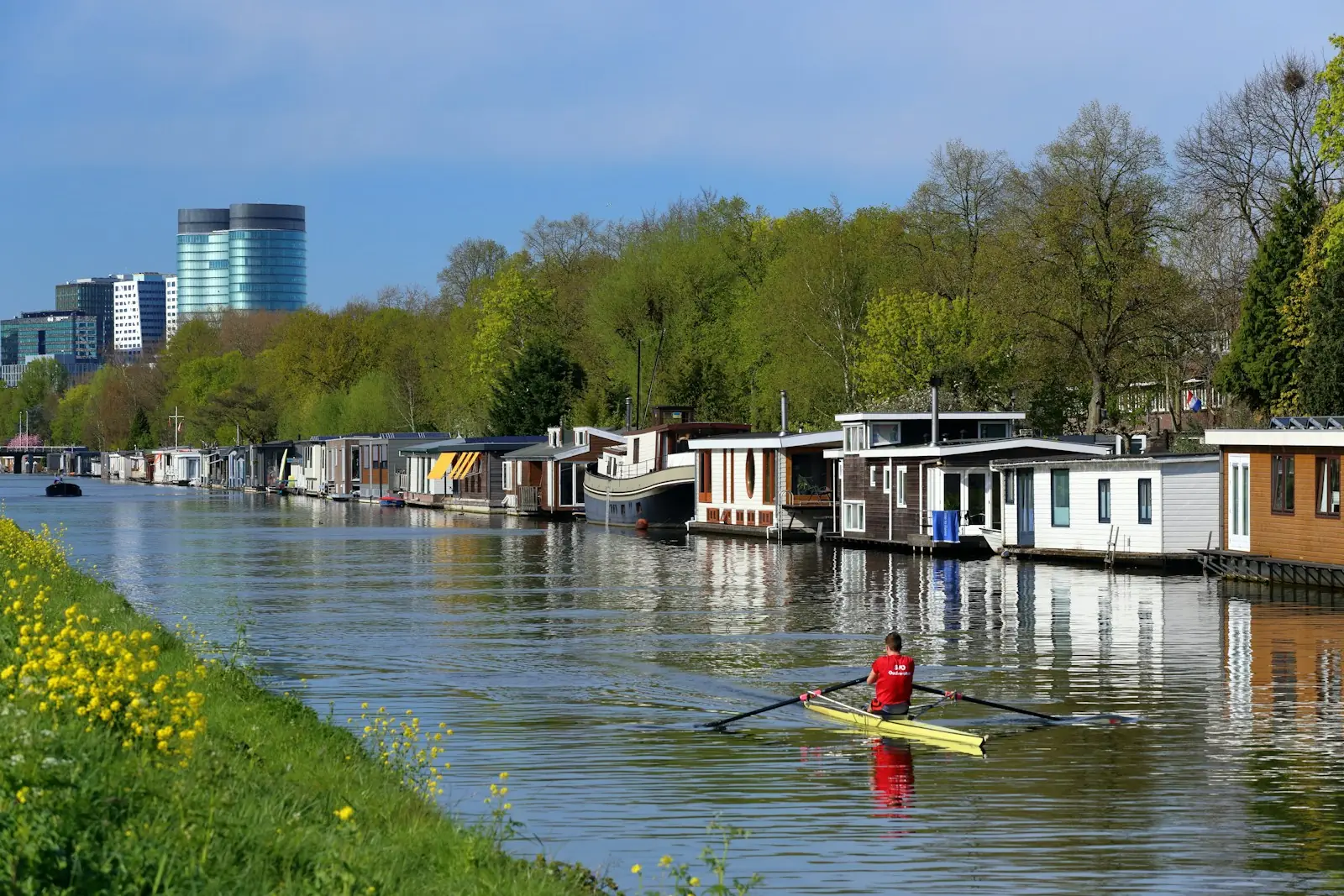 A single sculler on a calm river beside a row of houseboats, trees and town buildings beyond