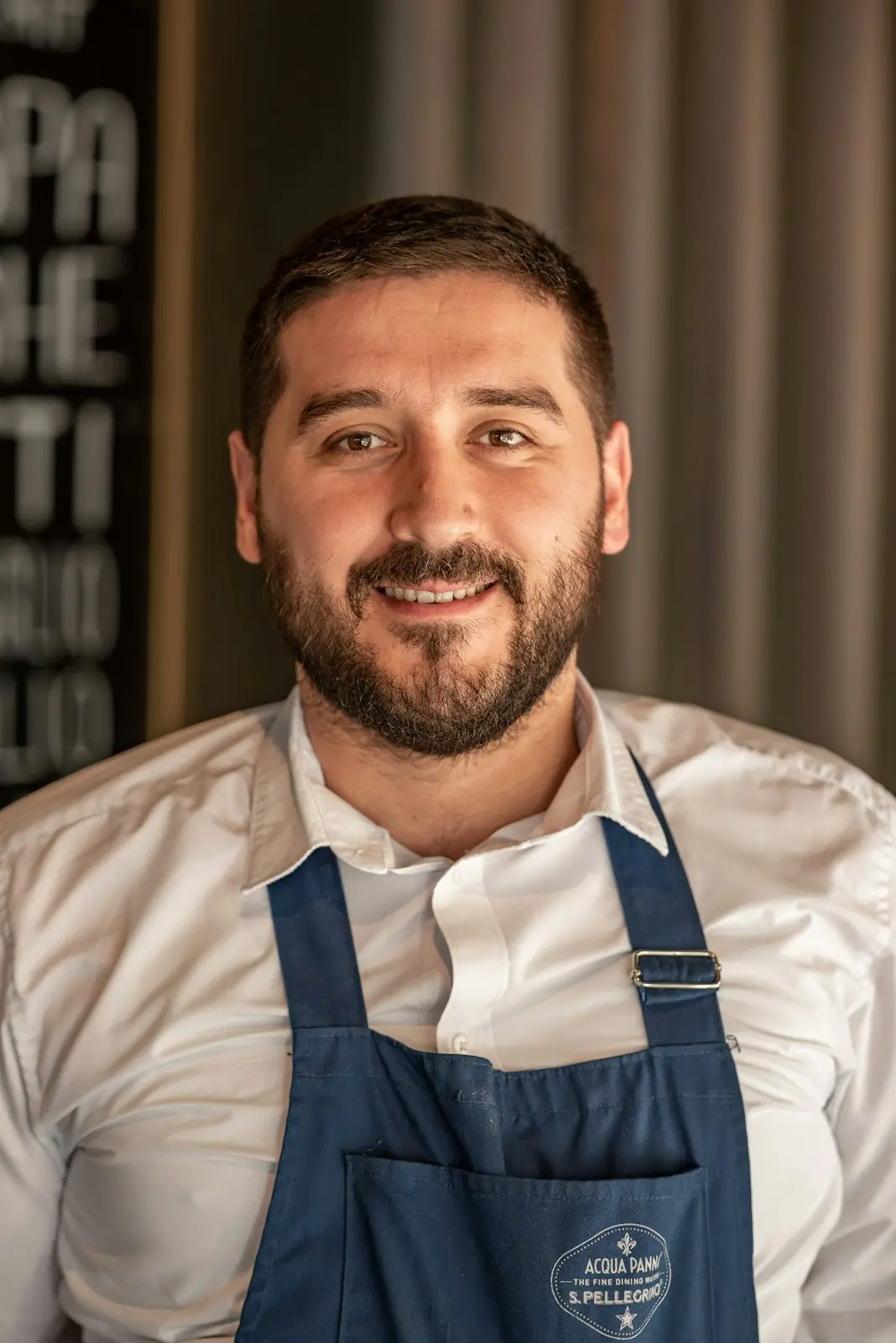 Portrait of Tom Fairley, smiling, with a dark beard and a blue apron over a white shirt