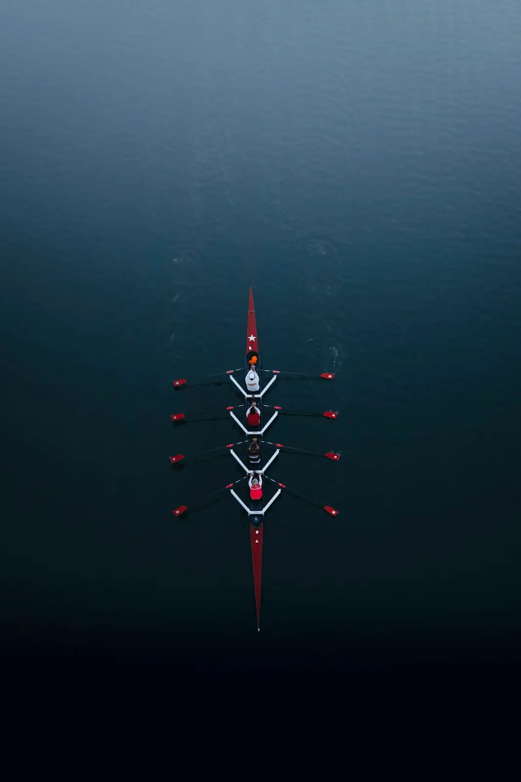 A coxed four from directly above on deep blue water, red-tipped oars fanned out in time
