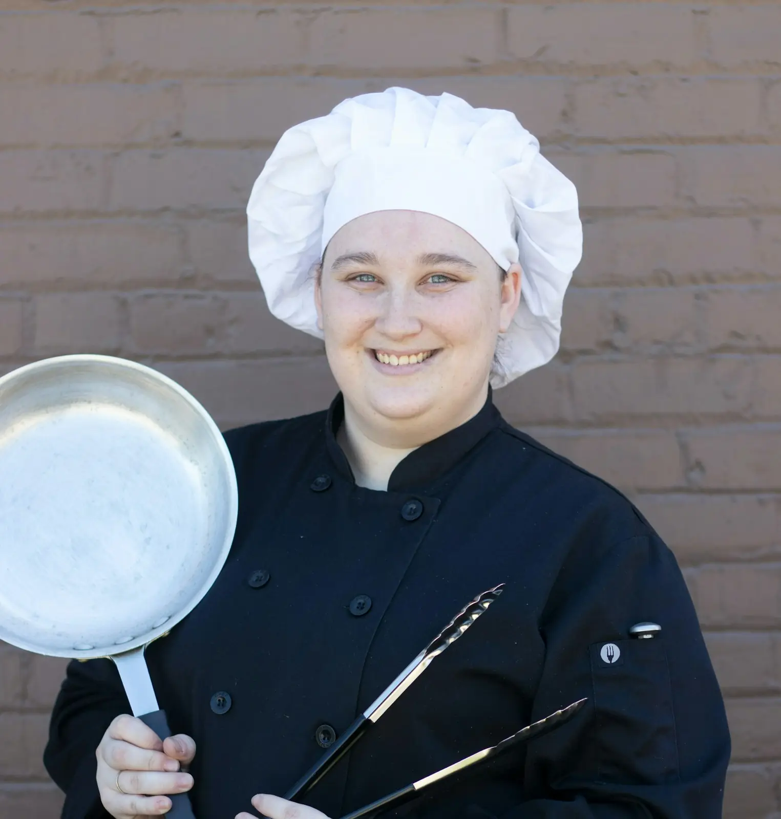 Portrait of Hanna Lindqvist in a white chef's hat and black jacket, smiling, holding a pan