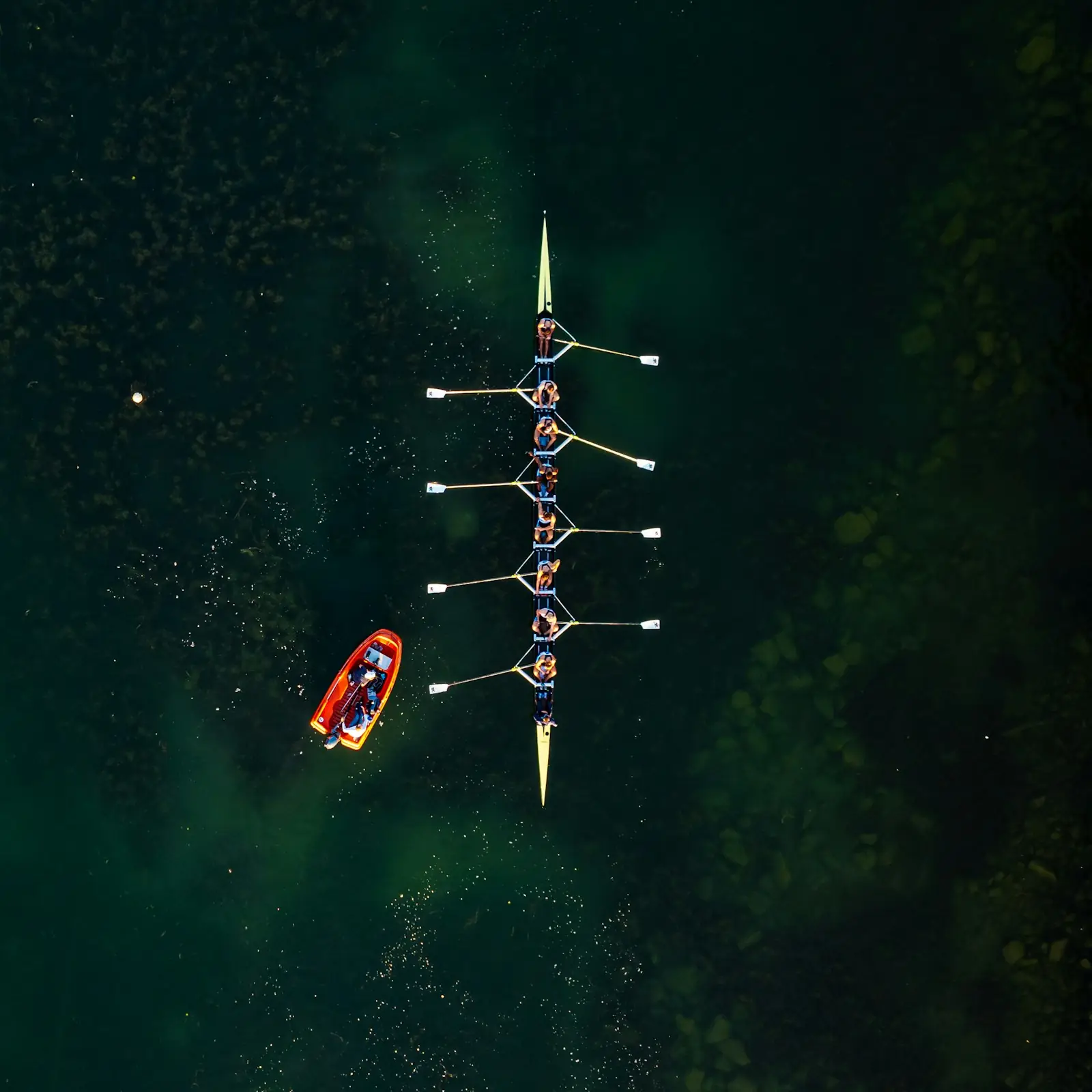 An eight from above on dark green water with a small red launch following alongside