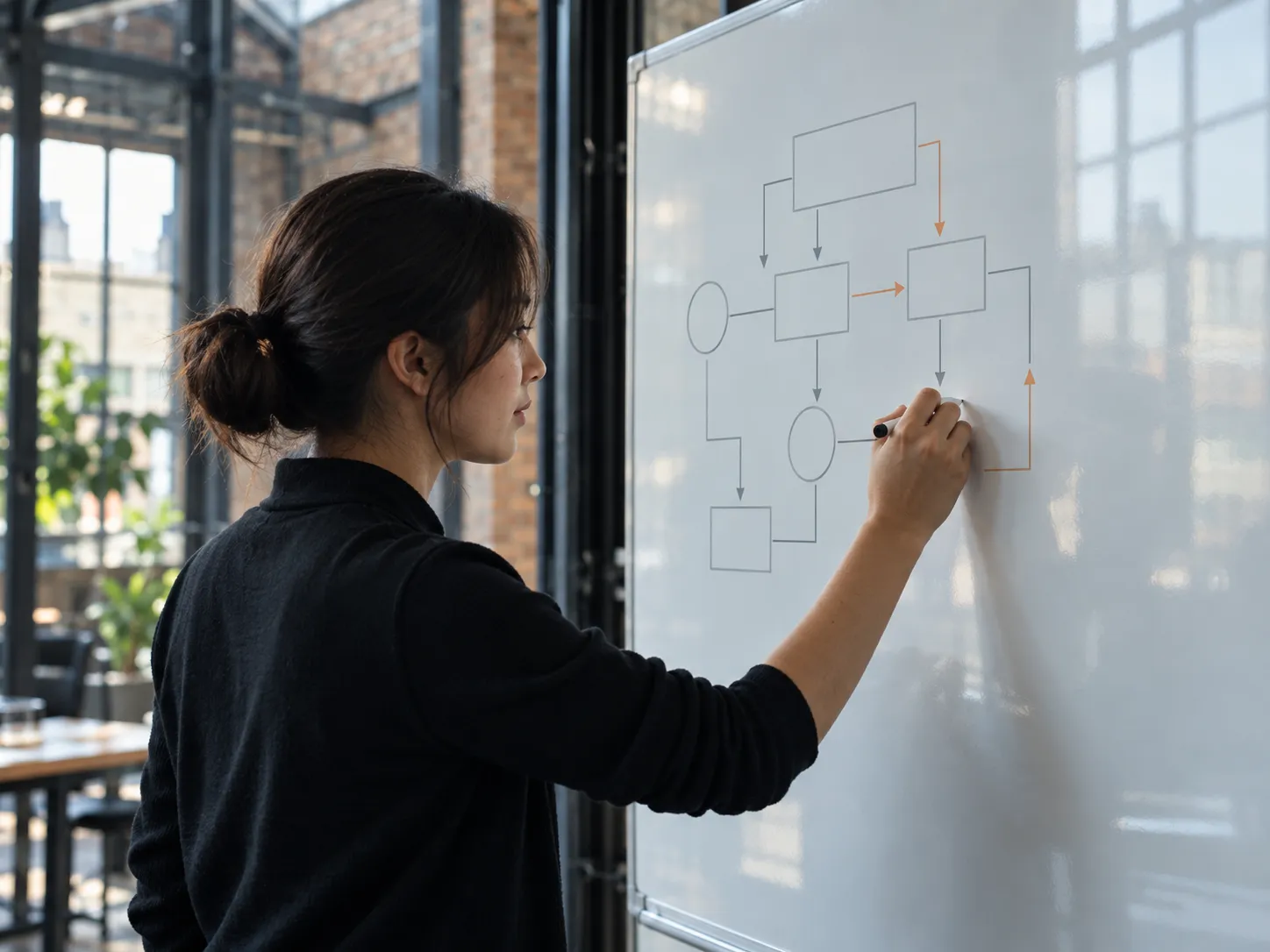 A woman draws boxes and arrows on a whiteboard beside a tall steel-framed window