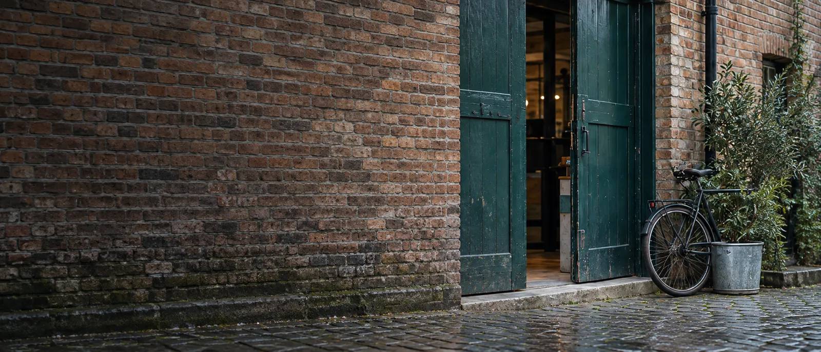 An open green timber loading door in a long brick wall, a bicycle and a potted olive beside it on wet cobbles