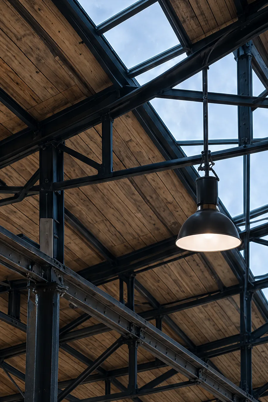 Timber roof trusses and steel north-light glazing seen from below, a black pendant lamp hanging in front