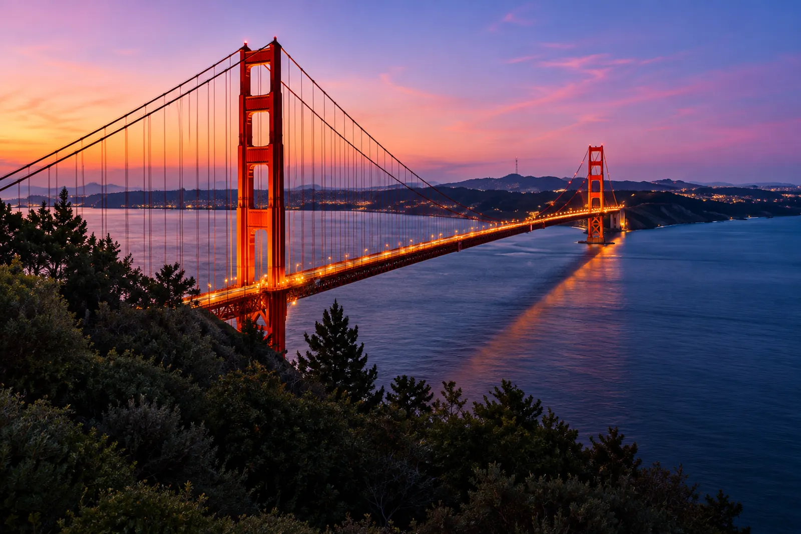 An orange suspension bridge across a bay at sunset, its lights glowing along the deck