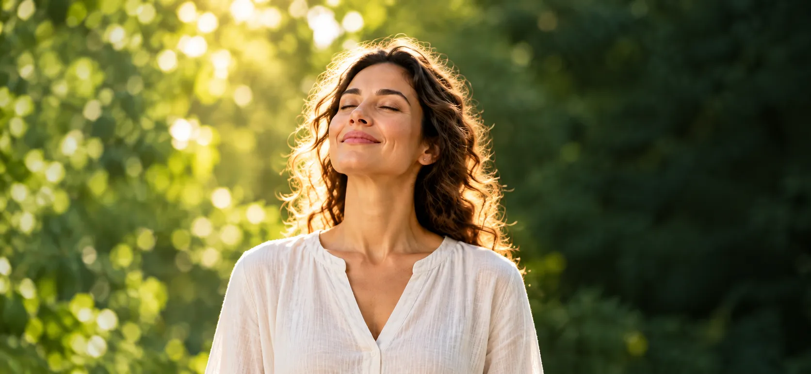 A woman with her eyes closed and her face lifted to the warm sunlight, among green trees