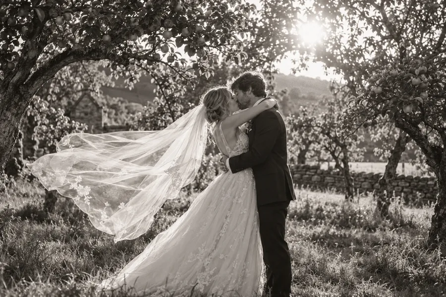 Black-and-white photograph of a couple kissing in an orchard as her long veil lifts in the wind