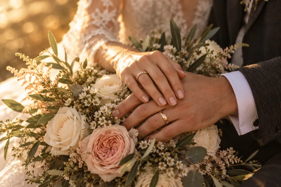 A couple's joined hands showing their wedding bands, resting on a bouquet of white and blush roses