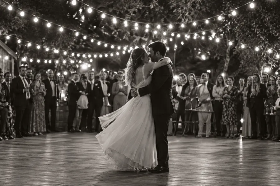 Black-and-white photograph of a couple's first dance under festoon lights, guests watching