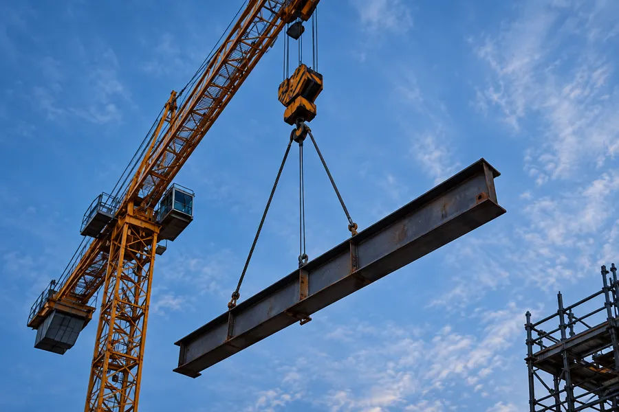 Looking up at a yellow crane hook lifting a steel I-beam against a blue sky with light cloud