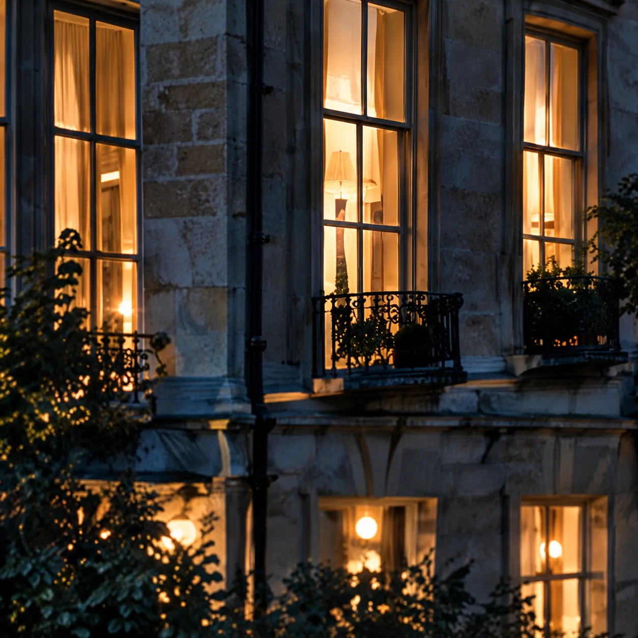 Tall lit sash windows in a stone building at evening, behind a tree