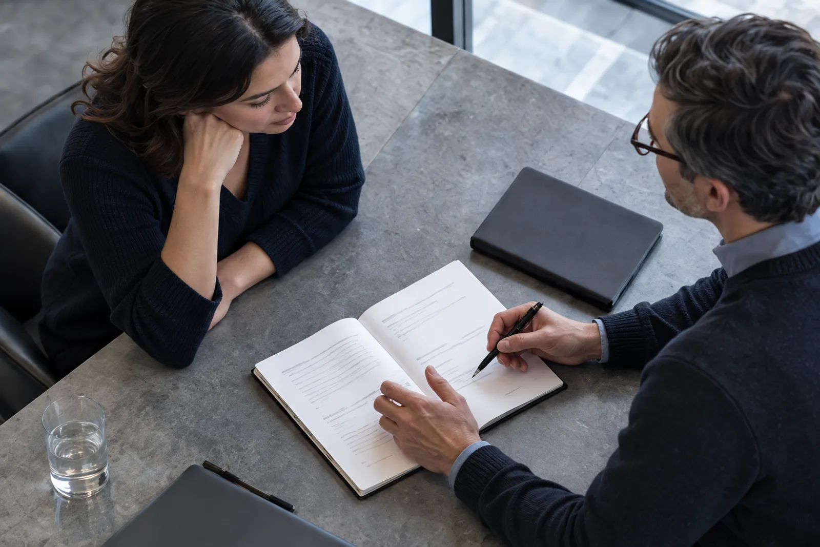 Seen from above, two people in dark knitwear at a grey stone table, a man writing in an open notebook while a woman leans in