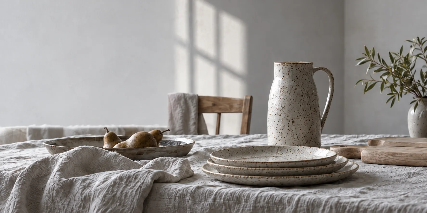 A dining table laid with grey linen, speckled plates, a tall jug, pale ash boards and a bowl of pears