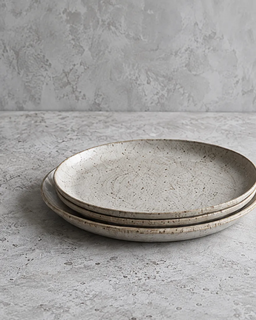 Three speckled oat-glazed stoneware dinner plates stacked on a pale surface