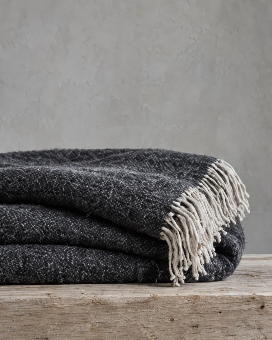 A folded charcoal lambswool throw with a fringed edge on a pale wooden plinth