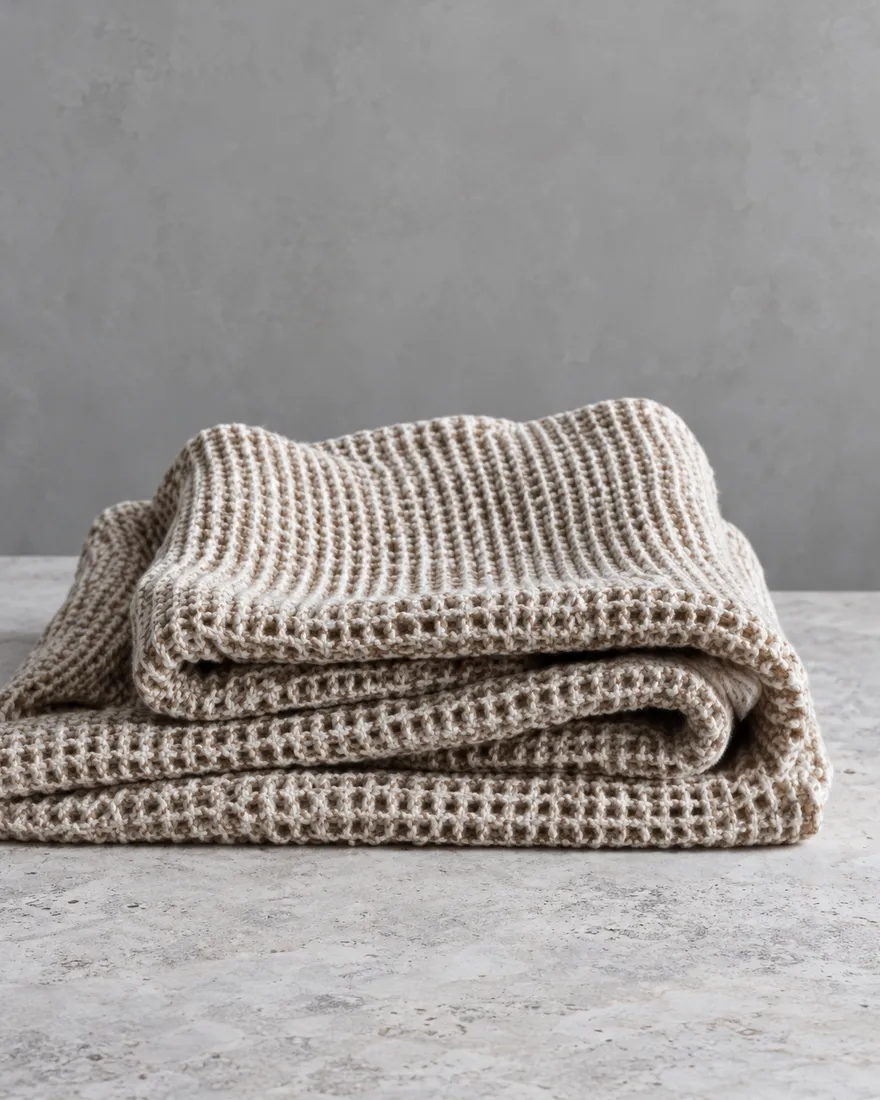A folded oat waffle cotton blanket on a pale stone surface