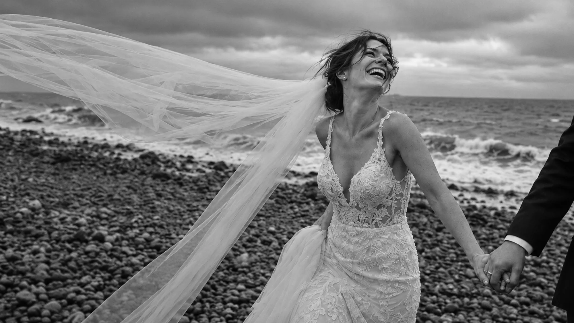 Bride laughing on a pebble beach as her long veil blows sideways, holding the groom's hand