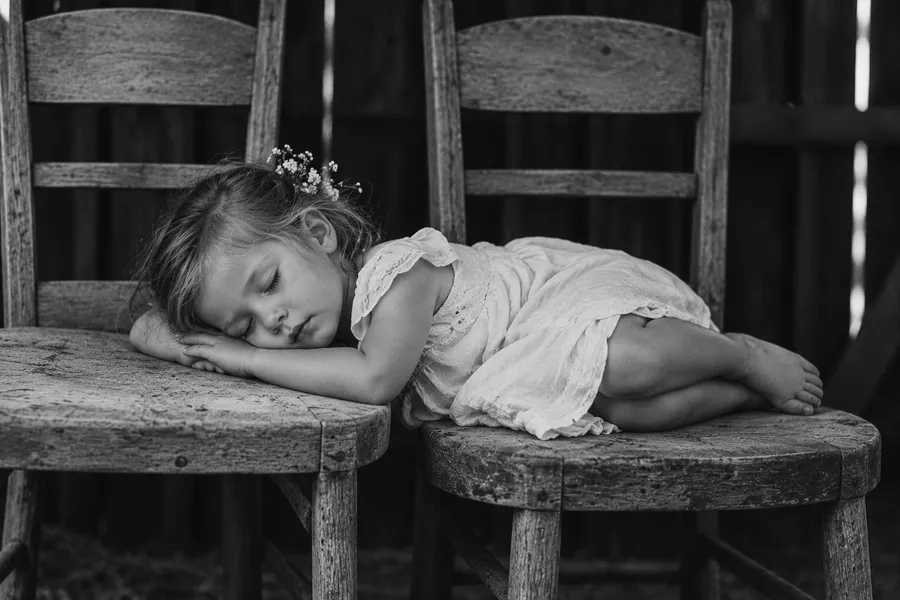 A little girl in a white dress asleep across two old wooden chairs