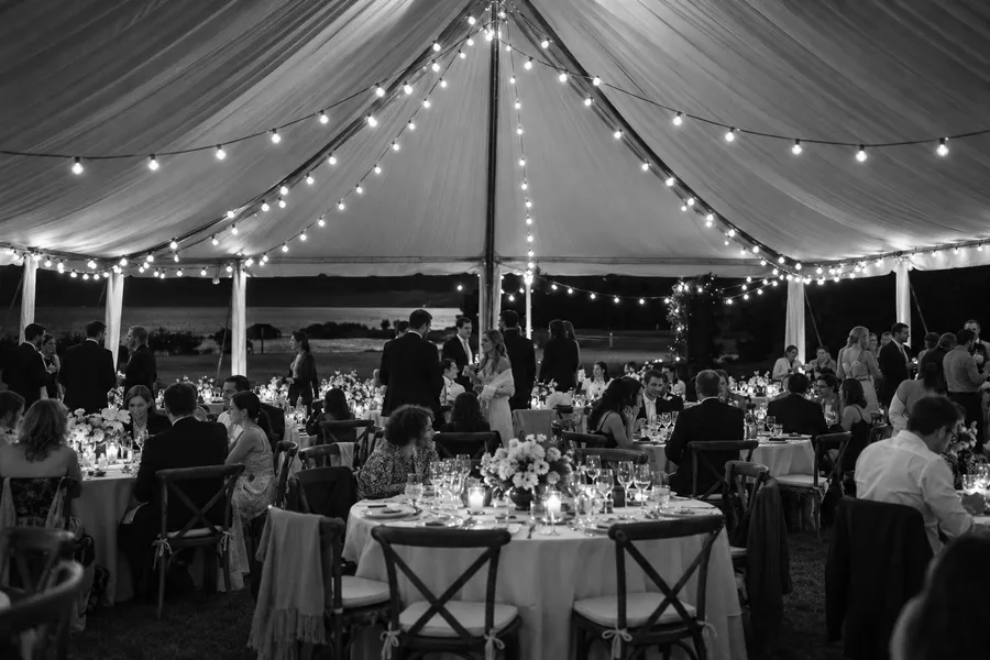 A sailcloth tent reception at night, bulb lights strung above round tables of guests