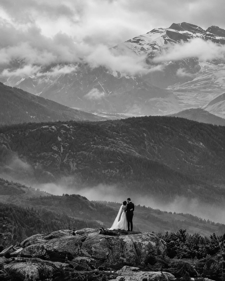 A small couple on a rocky ridge in a misty mountain valley under a snowy peak