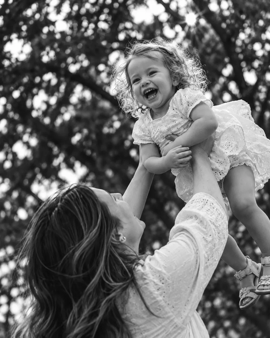 Mother lifting her laughing toddler into the air under trees