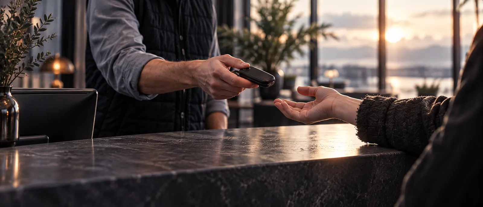 Across a dark counter an attendant’s hand offers a plain key fob to a traveller’s open hand in soft window light