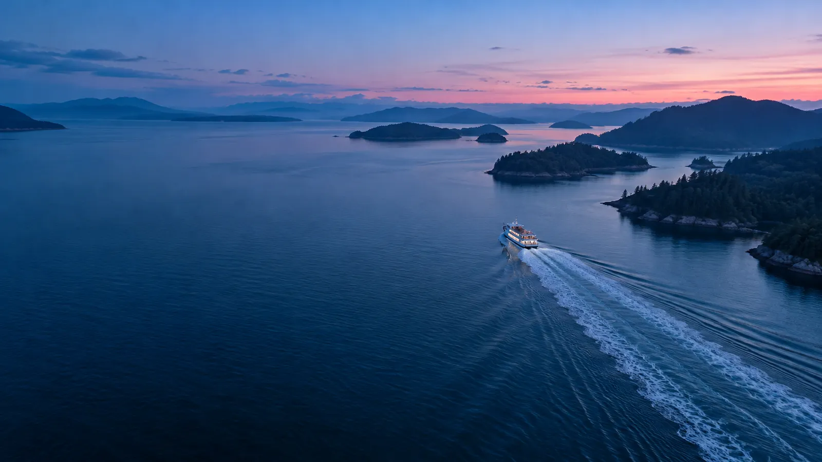 A blue-hour aerial of a small ferry drawing a long white wake through ink-blue water between dark islands