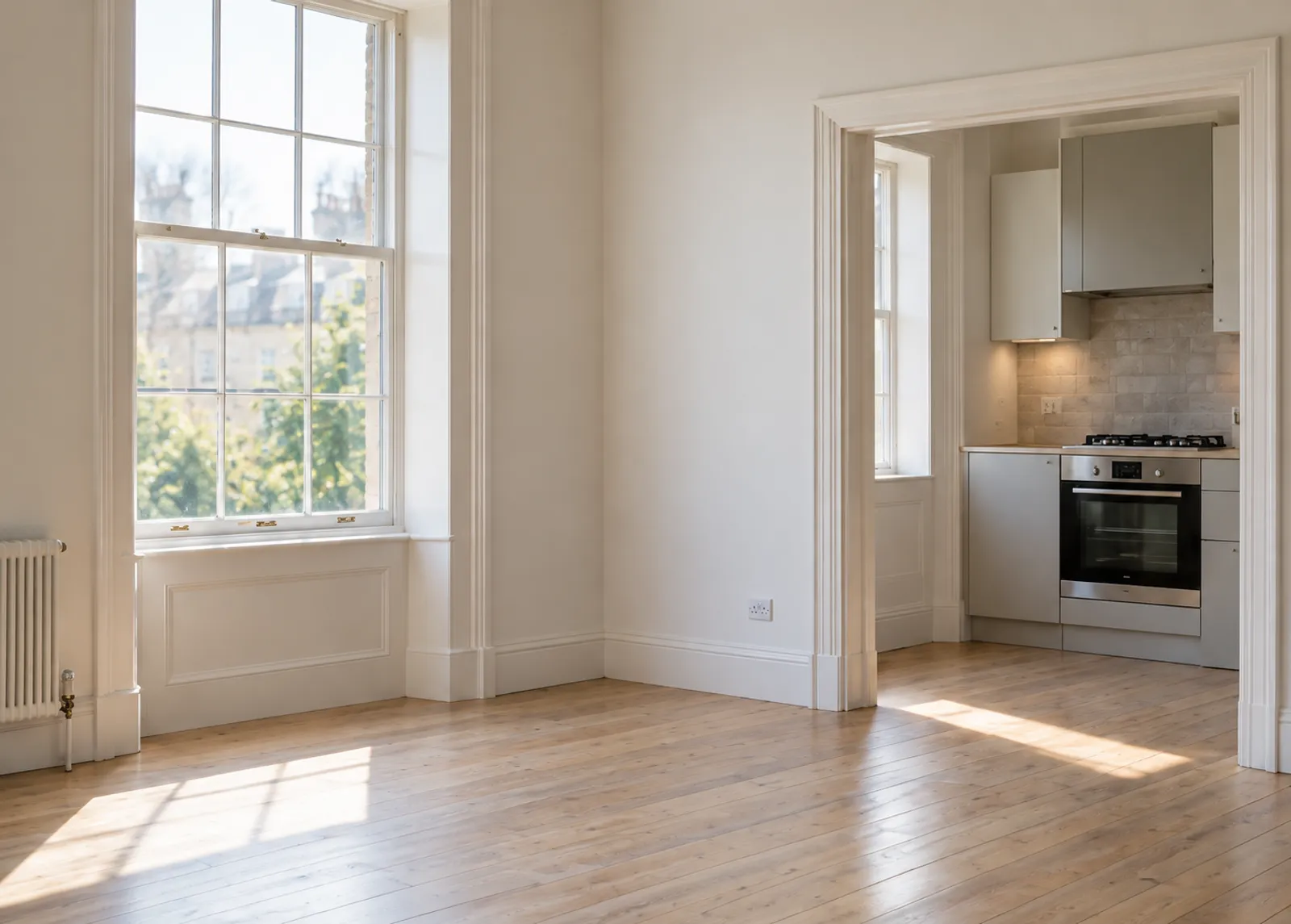 An empty, freshly cleaned flat: a white room with a tall sash window and pale floorboards, a clean kitchen and oven through the doorway