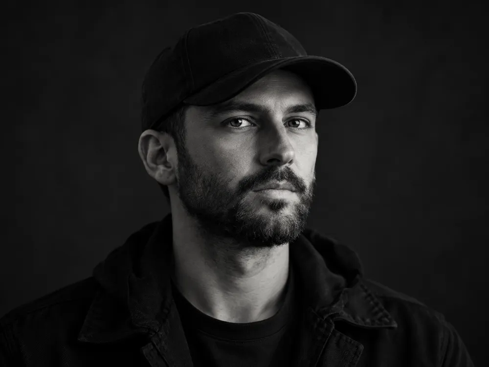 Black and white portrait of Noah Brandt, a bearded man in a dark cap and jacket