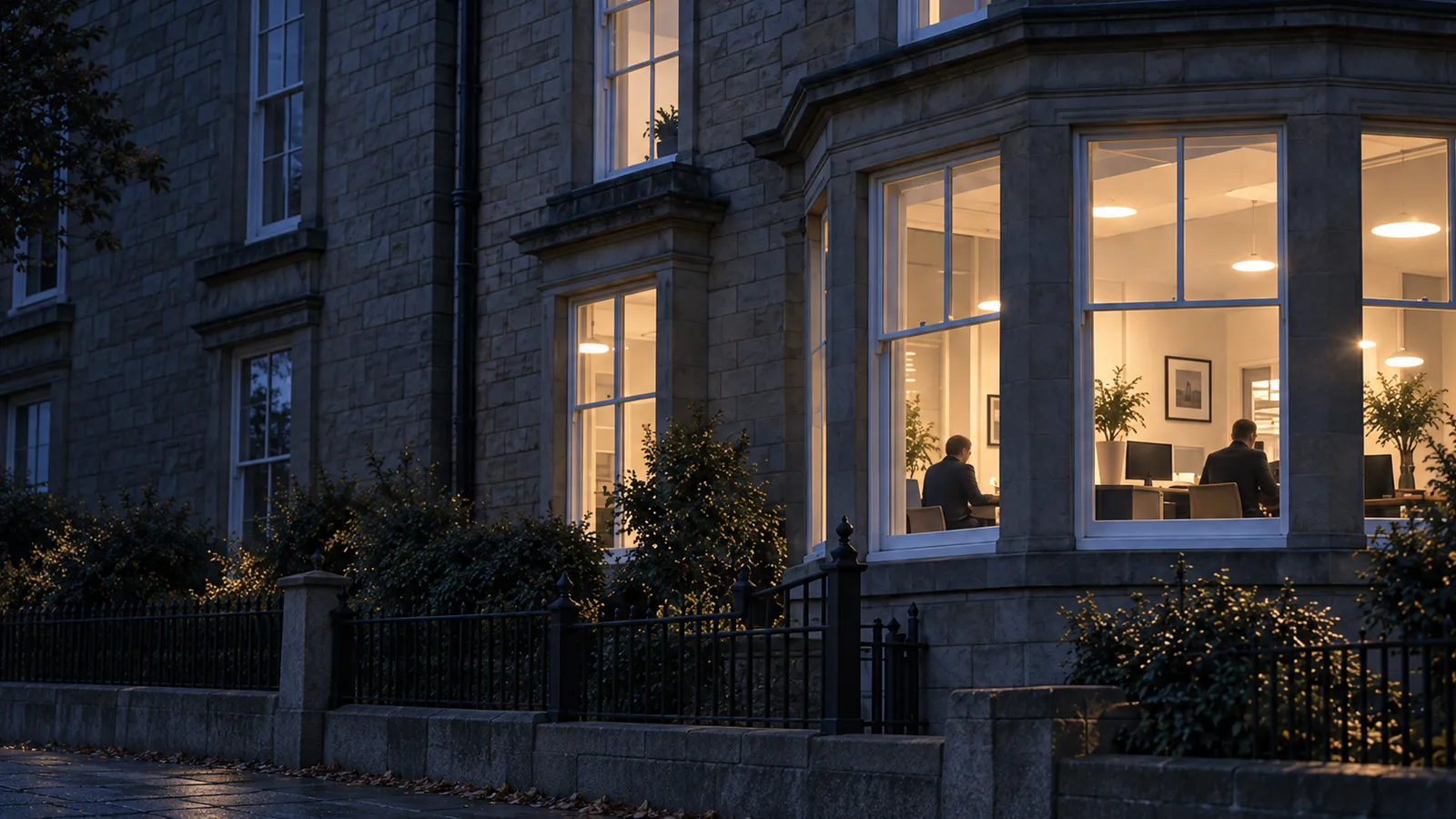 The lit bay windows of a first-floor office in a stone building at dusk, two people working at their desks inside